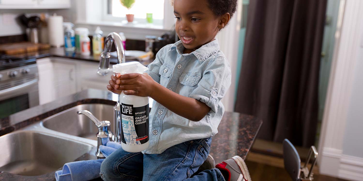 A super excited kid kneeling down on a granite countertop spraying Better Life's Stone and Granite Cleaner