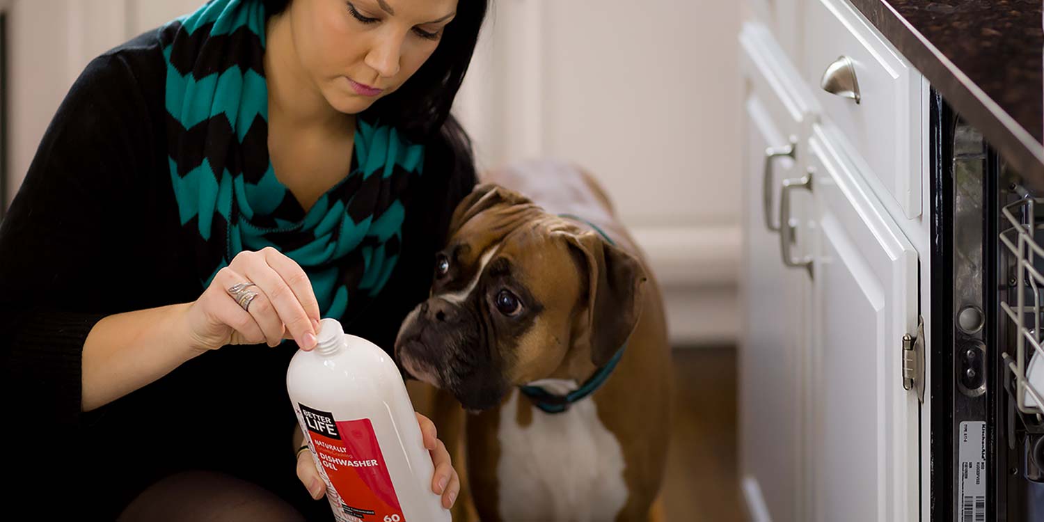 A woman opening up a bottle of Better Life's Dishwasher Cleaner with a puppy dog watching closely